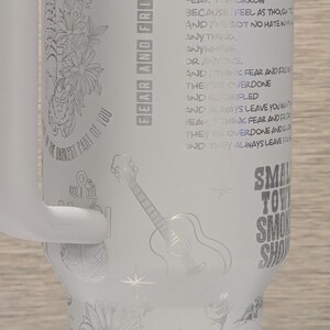 Może przedstawiać: Biały kubek termiczny z uchwytem, ​​z wytrawionymi wzorami kwiat&oacute;w, gitary i tekstu. Kubek zawiera słowa "Fear and Fridays" i cytat. Wytrawiony jest r&oacute;wnież słonecznik i kowboj.