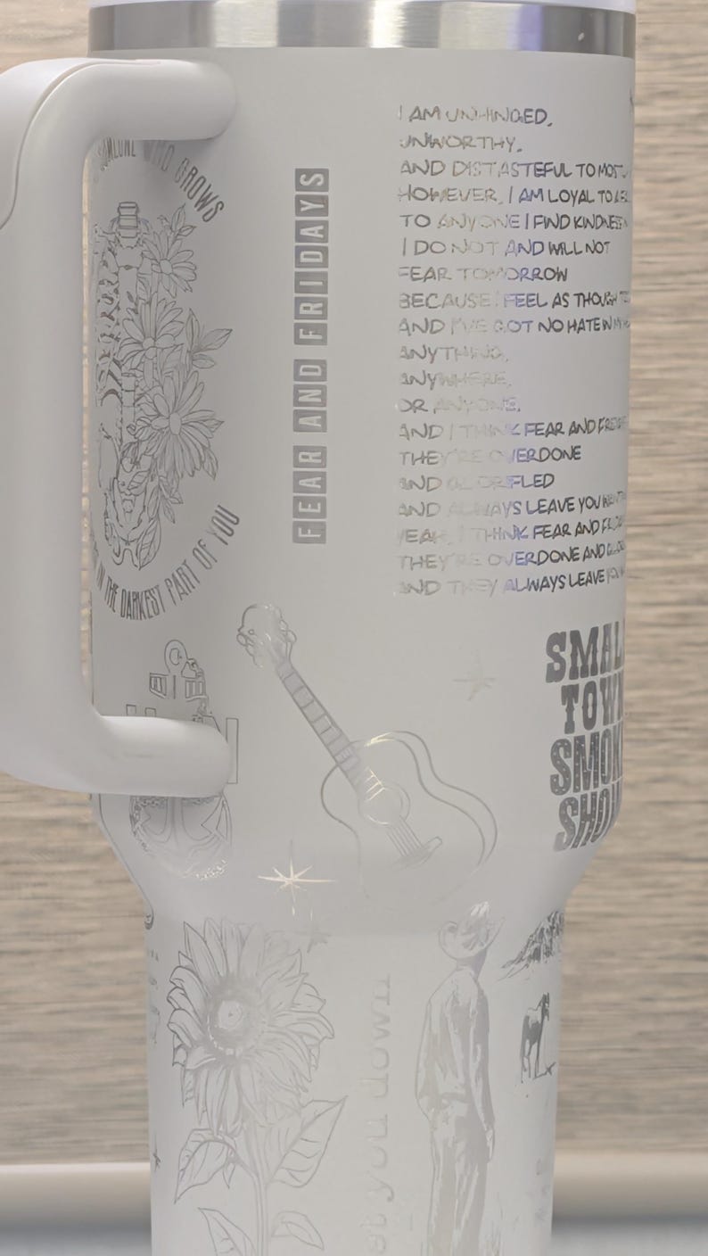 Może przedstawiać: Biały kubek termiczny z uchwytem, ​​z wytrawionymi wzorami kwiat&oacute;w, gitary i tekstu. Kubek zawiera słowa "Fear and Fridays" i cytat. Wytrawiony jest r&oacute;wnież słonecznik i kowboj.