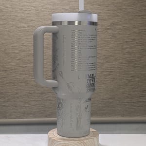 Może przedstawiać: Szary kubek termiczny z białą pokrywką i uchwytem. Kubek ma nadrukowany wz&oacute;r z tekstem, gitarą i słonecznikiem. Tekst zawiera "Small Town Smoke Show" i "let you down". Stoi na drewnianej podstawce.