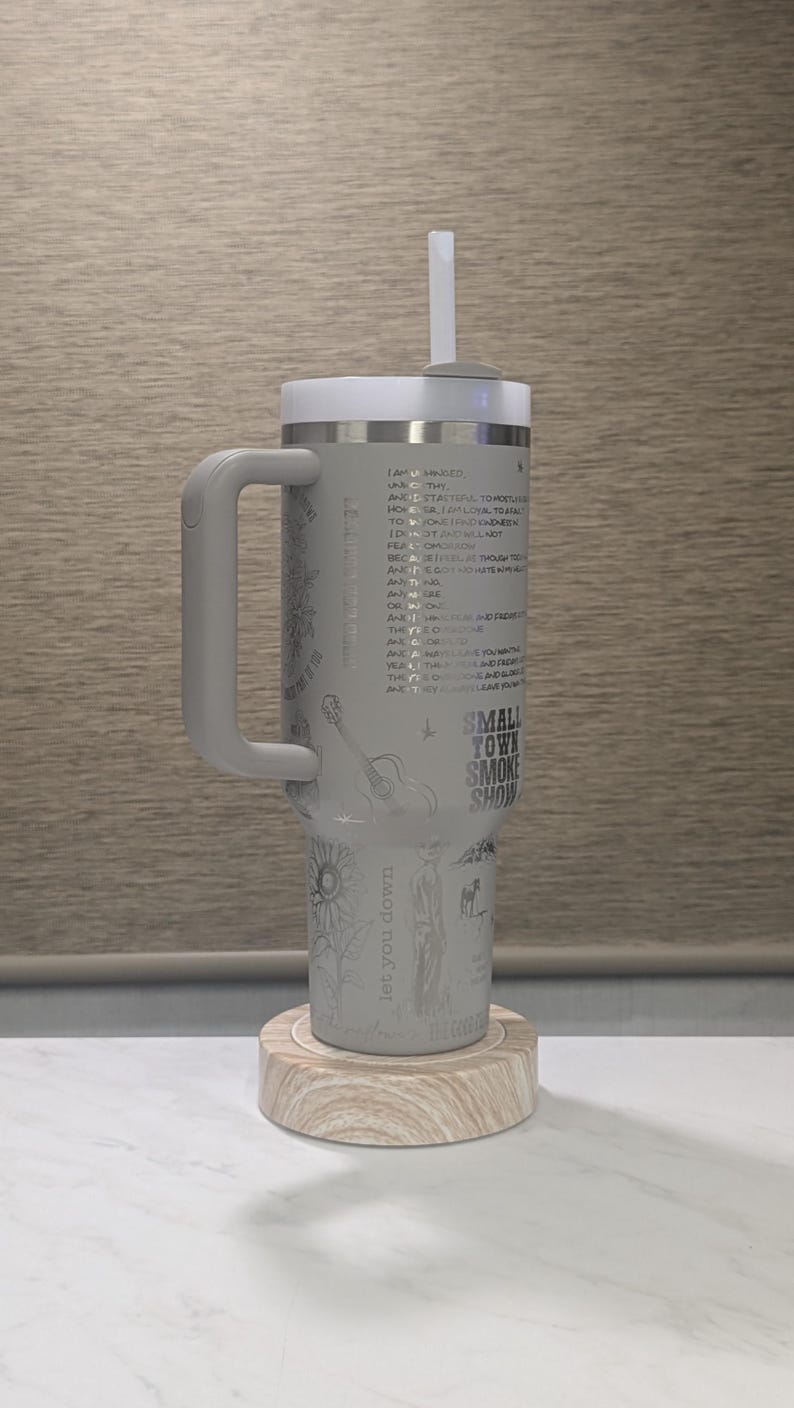 Może przedstawiać: Szary kubek termiczny z białą pokrywką i uchwytem. Kubek ma nadrukowany wz&oacute;r z tekstem, gitarą i słonecznikiem. Tekst zawiera "Small Town Smoke Show" i "let you down". Stoi na drewnianej podstawce.