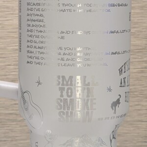 Może przedstawiać: Biały kubek ze srebrnym tekstem i ilustracjami. Zawiera tekst "Small Town Smoke Show" i inne frazy. Zawiera obrazy gitary, kowboja, konia i innych wzor&oacute;w.