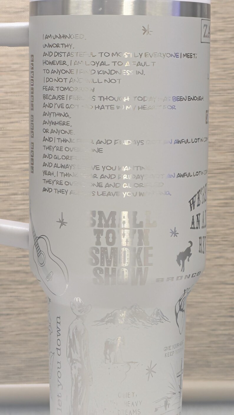 Może przedstawiać: Biały kubek ze srebrnym tekstem i ilustracjami. Zawiera tekst "Small Town Smoke Show" i inne frazy. Zawiera obrazy gitary, kowboja, konia i innych wzor&oacute;w.