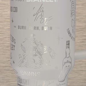 Może przedstawiać: Biały kubek Stanley z wytrawionymi wzorami i tekstem. Kubek zawiera słowa "BRYAN", "tourniquet", "STANLEY" i "JIM BEAM". Inne wytrawione teksty to "THE BURN, BURN, BURN", "STRAIGHT OUTTA OKLAHOMA" i "Baptize me in a bottle of Beam".