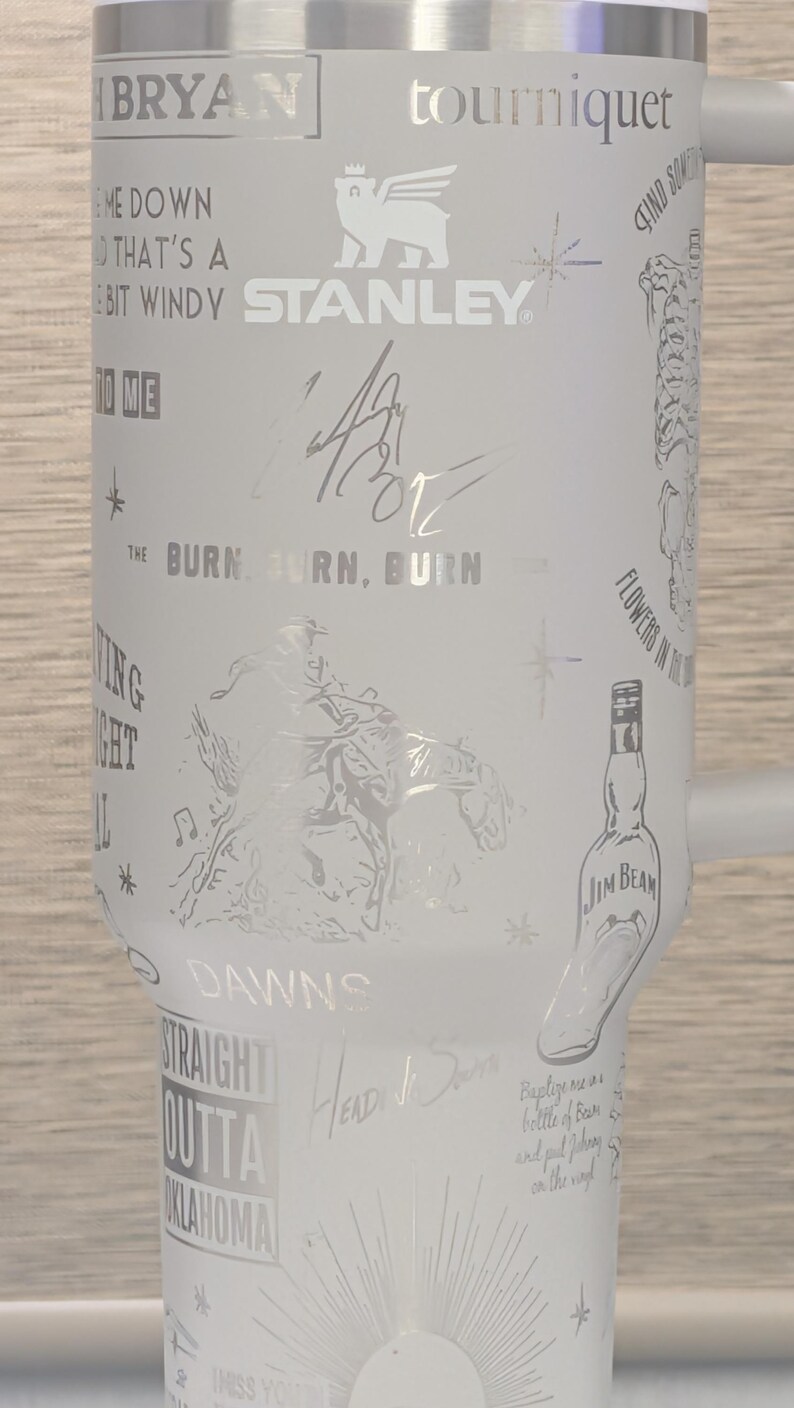 Może przedstawiać: Biały kubek Stanley z wytrawionymi wzorami i tekstem. Kubek zawiera słowa "BRYAN", "tourniquet", "STANLEY" i "JIM BEAM". Inne wytrawione teksty to "THE BURN, BURN, BURN", "STRAIGHT OUTTA OKLAHOMA" i "Baptize me in a bottle of Beam".