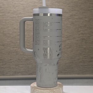 Może przedstawiać: Szary kubek z uchwytem, białą pokrywką i słomką. Kubek posiada wytrawiony tekst i ilustracje, w tym słowa "Small Town Smoke Show". Spoczywa na drewnianym spodku.