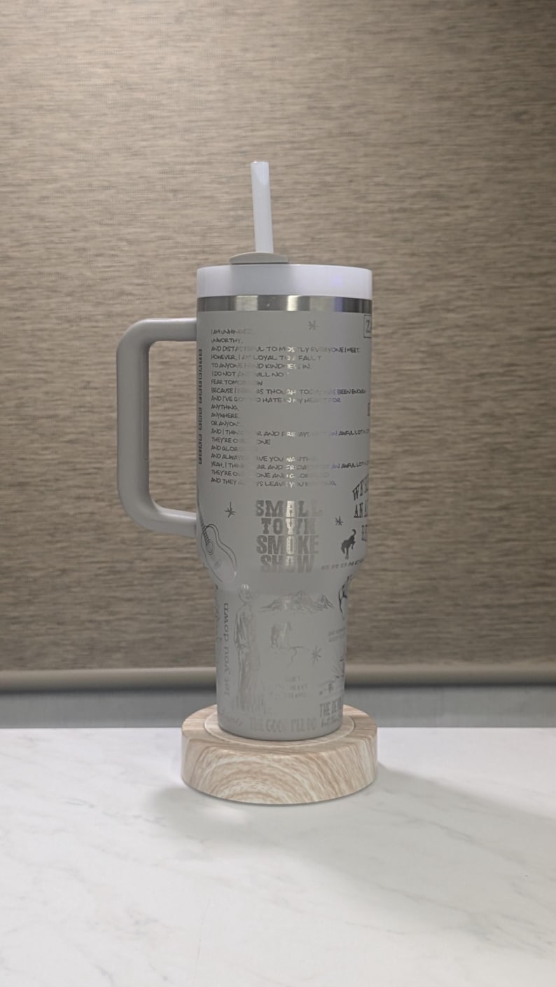 Może przedstawiać: Szary kubek z uchwytem, białą pokrywką i słomką. Kubek posiada wytrawiony tekst i ilustracje, w tym słowa "Small Town Smoke Show". Spoczywa na drewnianym spodku.