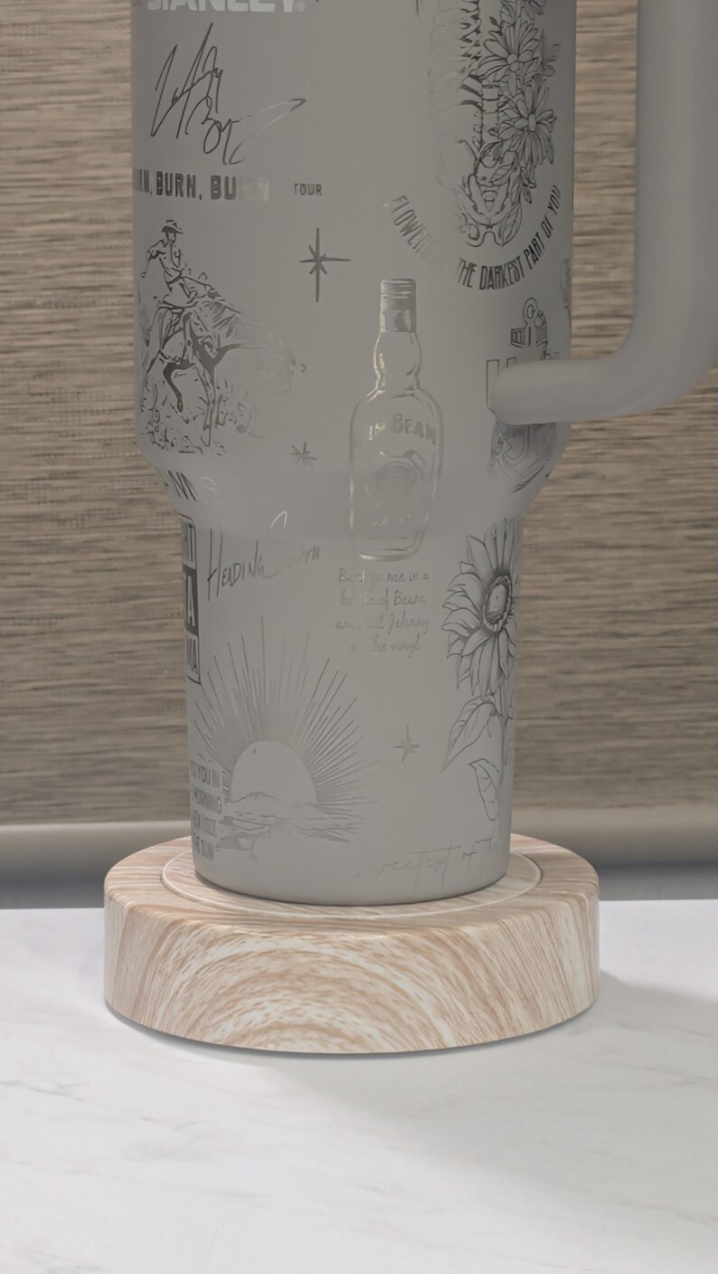 Może przedstawiać: Biały, izolowany kubek z wytrawionymi ilustracjami kowboja, kwiat&oacute;w, butelki Jim Beam i wschodzącego słońca. Kubek stoi na okrągłym, drewnianym spodku. Kubek ma uchwyt.