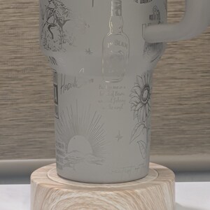 Może przedstawiać: Jasnoszary kubek Stanley z wytrawionymi ilustracjami kowboja, kwiat&oacute;w, butelki Jim Beam i wschodu słońca. Kubek stoi na okrągłym drewnianym spodku.