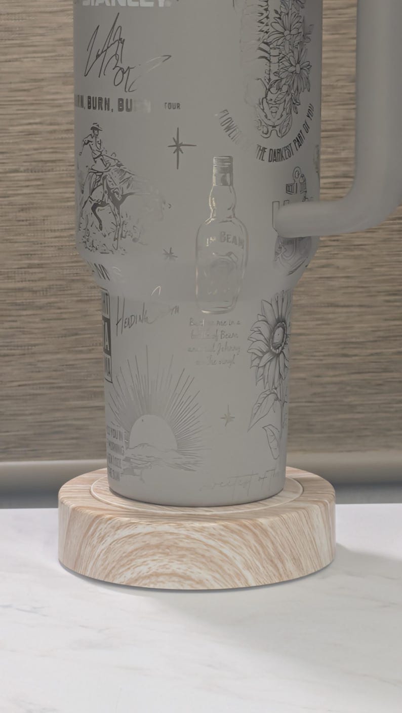 Może przedstawiać: Jasnoszary kubek Stanley z wytrawionymi ilustracjami kowboja, kwiat&oacute;w, butelki Jim Beam i wschodu słońca. Kubek stoi na okrągłym drewnianym spodku.