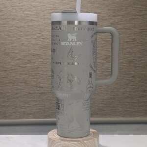 Może przedstawiać: Szary kubek Stanley z uchwytem, słomką i pokrywką, spoczywający na drewnianej podstawce. Kubek jest wytrawiony r&oacute;żnymi wzorami, w tym tekstem, podpisami i ilustracjami. Kubek ma około 25 cm wysokości.