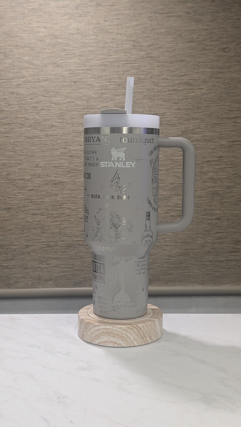 Może przedstawiać: Szary kubek Stanley z uchwytem, słomką i pokrywką, spoczywający na drewnianej podstawce. Kubek jest wytrawiony r&oacute;żnymi wzorami, w tym tekstem, podpisami i ilustracjami. Kubek ma około 25 cm wysokości.