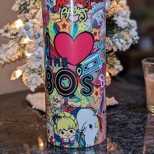 May include: A stainless steel tumbler with a colorful design featuring 80s pop culture icons like My Little Pony, Care Bears, and the Teenage Mutant Ninja Turtles. The tumbler has a heart with the text "I Love the 80s" and a rainbow.