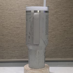 Może przedstawiać: Szary kubek termiczny ze stali nierdzewnej z białą pokrywką i słomką. Kubek ma wytrawione wzory, w tym słonecznik i gitarę. Stoi na drewnianej podstawce.