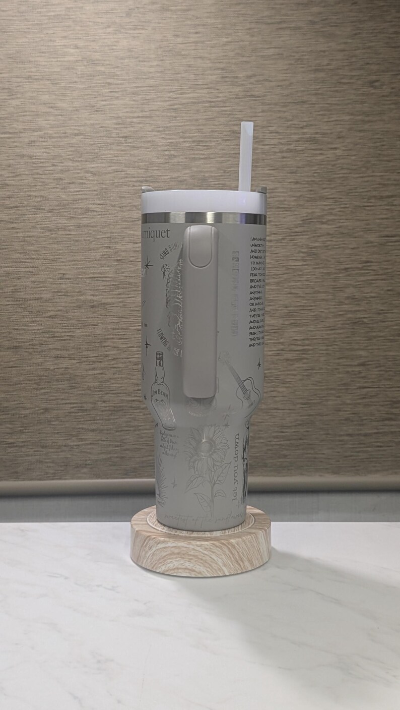 Może przedstawiać: Szary kubek termiczny ze stali nierdzewnej z białą pokrywką i słomką. Kubek ma wytrawione wzory, w tym słonecznik i gitarę. Stoi na drewnianej podstawce.