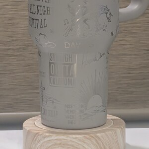 Może przedstawiać: Jasnoszary kubek z wytrawionymi wzorami i tekstem, w tym "Burn, Burn, Burn" i "Straight Outta Oklahoma". Kubek stoi na jasnej drewnianej podstawce. Uchwyt jest jasnoszary.