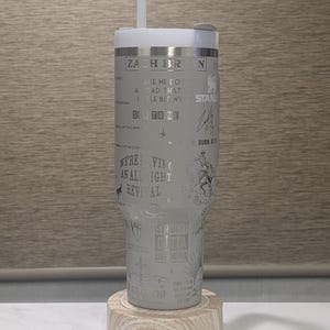 Może przedstawiać: Biały kubek ze słomką, z wytrawionym tekstem i ilustracjami. Projekt zawiera tekst "ZACH BR", "WE'RE VING AN ALL IGHT REVIVAL" i inne frazy. Spoczywa na drewnianej podstawce.