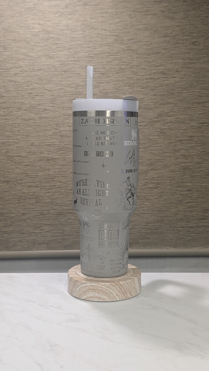 Może przedstawiać: Biały kubek ze słomką, z wytrawionym tekstem i ilustracjami. Projekt zawiera tekst "ZACH BR", "WE'RE VING AN ALL IGHT REVIVAL" i inne frazy. Spoczywa na drewnianej podstawce.