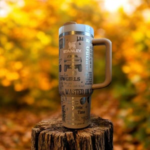 Puede incluir: Una taza de viaje de acero inoxidable marrón con un asa negra. La taza tiene una variedad de texto y gráficos dorados, incluyendo las palabras "Cowgirls", "Outlaw", "Wasted on You" y "Quittin' Time". La taza está sentada sobre un tocón de árbol frente a un fondo borroso de follaje otoñal.