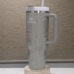 Może przedstawiać: Szary kubek termiczny z uchwytem, pokrywką i słomką. Kubek pokryty jest wytrawionymi wzorami i tekstem, w tym logo "STANLEY". Stoi na drewnianej podstawce.