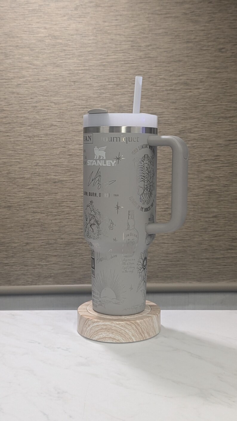 Może przedstawiać: Szary kubek termiczny z uchwytem, pokrywką i słomką. Kubek pokryty jest wytrawionymi wzorami i tekstem, w tym logo "STANLEY". Stoi na drewnianej podstawce.