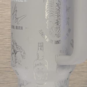 Może przedstawiać: Biały kubek termiczny z wytrawionymi wzorami, w tym tekstem, szkieletem, kwiatami, butelką Jim Beam i słonecznikiem. Posiada uchwyt i logo Stanley. Przeznaczony do napoj&oacute;w.