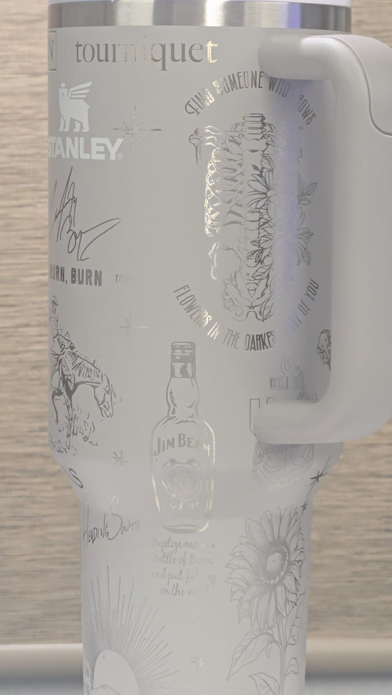 Może przedstawiać: Biały kubek termiczny z wytrawionymi wzorami, w tym tekstem, szkieletem, kwiatami, butelką Jim Beam i słonecznikiem. Posiada uchwyt i logo Stanley. Przeznaczony do napoj&oacute;w.