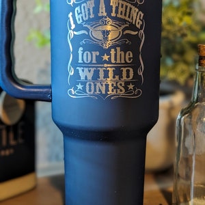 May include: A dark blue stainless steel travel mug with a black handle. The mug has a silver inscription that reads "I got a thing for the wild ones" with a star and a stylized steer head.