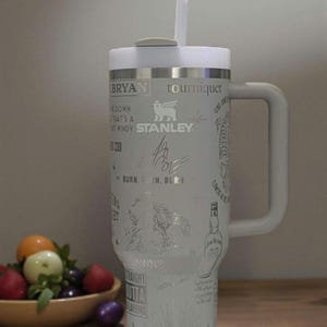 Może przedstawiać: Biały kubek Stanley z uchwytem i słomką. Kubek posiada grawerowane wzory i tekst, w tym słowo "Stanley" i tekst "BRYAN tourniquet". Kubek stoi na drewnianej powierzchni.