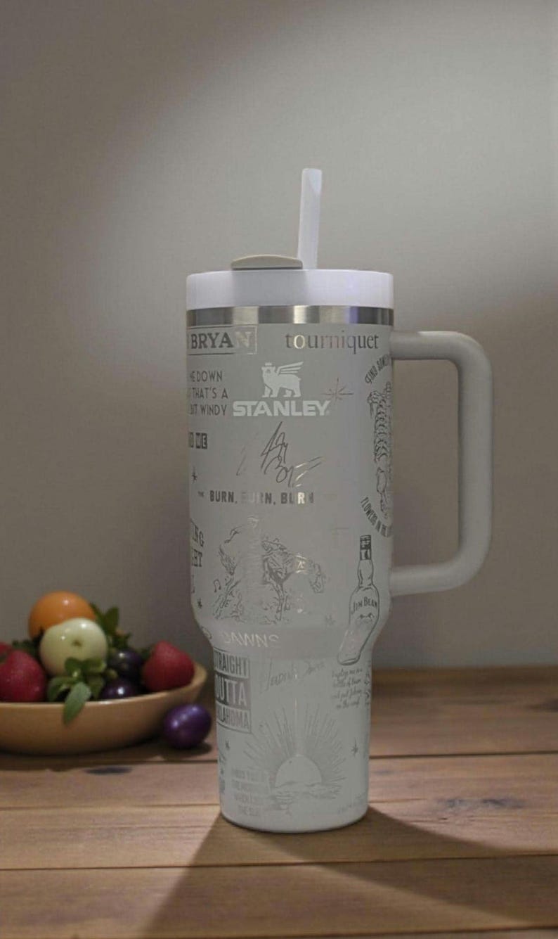 Może przedstawiać: Biały kubek Stanley z uchwytem i słomką. Kubek posiada grawerowane wzory i tekst, w tym słowo "Stanley" i tekst "BRYAN tourniquet". Kubek stoi na drewnianej powierzchni.