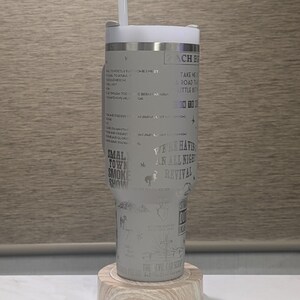 Może przedstawiać: Jasnoszary kubek z białą pokrywką i słomką, spoczywający na drewnianej podstawce. Kubek zawiera wytrawiony tekst i ilustracje, w tym słowa "Small Town Smoke Show" i "We're Having An All Night Revival."