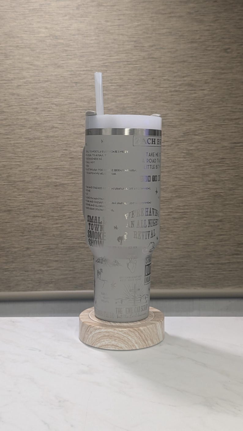 Może przedstawiać: Jasnoszary kubek z białą pokrywką i słomką, spoczywający na drewnianej podstawce. Kubek zawiera wytrawiony tekst i ilustracje, w tym słowa "Small Town Smoke Show" i "We're Having An All Night Revival."