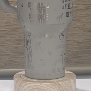 Może przedstawiać: Biały kubek z wytrawionymi ilustracjami i tekstem, w tym słowami "Small Town Smoke" i "We're an Alley Review". Kubek stoi na jasnej drewnianej podstawce.