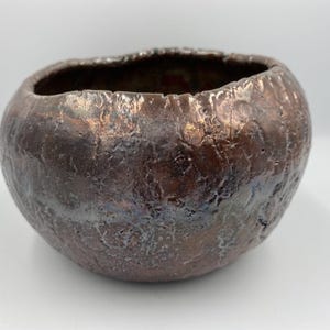 Könnte beinhalten: Eine handgefertigte, strukturierte Keramikschale mit dunklem, metallischem Finish. Die Schale hat eine abgerundete Form mit unebenem Rand und zeigt eine Mischung aus Braun-, Bronze- und Silbertönen. Dieses dekorative Stück eignet sich für die Inneneinrichtung.