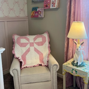 Puede incluir: Un sillón blanco con un cojín acolchado rosa y blanco. El sillón está en una guardería con papel tapiz rosa y blanco y una cortina de terciopelo rosa. Una mesa pequeña con una lámpara de conejo blanco está al lado de la silla.