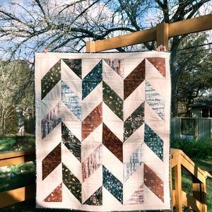 Puede incluir: Un edredón hecho a mano con un patrón de chevrón. El edredón presenta filas alternas de telas crema, marrón, verde y azul. El edredón se exhibe al aire libre, sostenido contra un fondo de cielo azul y ramas de árboles.