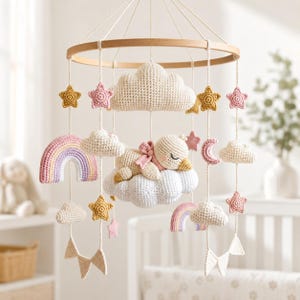May include: A nursery mobile featuring a circular wooden frame with hanging crocheted elements including a sleeping bird on a cloud, rainbows, stars, a crescent moon, and small clouds in pastel shades of pink, yellow, and white suspended by white cords.