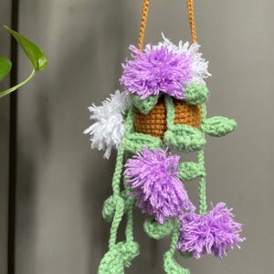 May include: A handmade crochet hanging planter with a brown pot, green leaves, and purple and white yarn flowers. It hangs from a braided orange cord. The design is whimsical and decorative, suitable for indoor use.