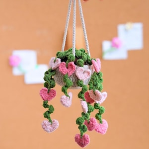May include: A handmade crochet hanging planter with a gray pot, filled with green crochet foliage. Pink and red heart-shaped crochet vines cascade down. The planter is suspended by gray cords.