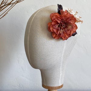 May include: A burnt orange fabric flower hair clip with green and brown accents. The flower is on a white mannequin head.