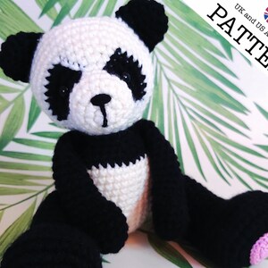 May include: A crocheted panda bear toy with black and white fur. The panda has a pink nose and pink paw pads. The toy is sitting on a green and white background.