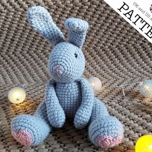 May include: A crocheted gray bunny rabbit with pink ears and a pink nose. The bunny is sitting on a brown textured surface with string lights.