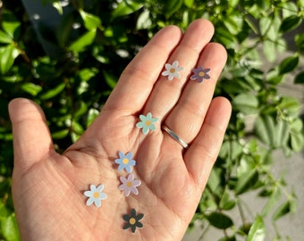 May include: A hand holding seven small daisy-shaped earrings with different coloured centres and petals. The earrings are made of metal and have a shiny finish.