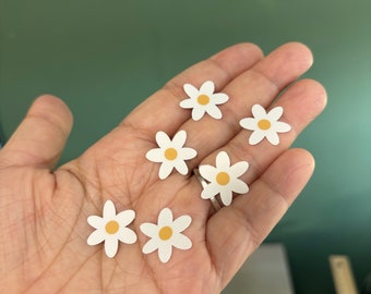 May include: A hand holding six white daisy stickers with yellow centres. The stickers are small and perfect for adding a touch of whimsy to crafts, journals, or other projects.