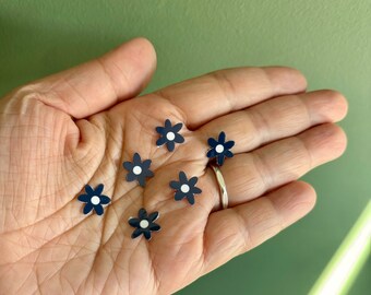 May include: Six small navy blue flower-shaped embellishments with white centres. The embellishments are on a person's hand.