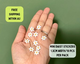 May include: A pack of 10 white daisy stickers with yellow centres. Each sticker is 1.3 cm wide. The stickers are on a hand.