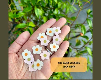 May include: A hand holding a variety of small daisy stickers. The stickers are pink, white and yellow with orange centres. The stickers are 1.6 cm wide. The text "MINI DAISY STICKERS 1.6CM WIDTH" is printed on a yellow rectangle.