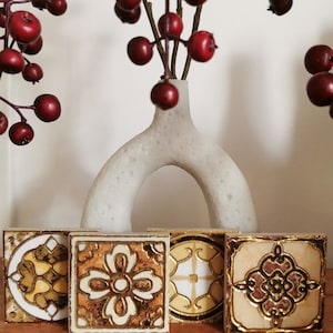 May include: Four square tiles featuring intricate gold and cream-colored decorative patterns. Each tile displays a unique geometric or floral design with metallic gold accents and raised textures, arranged in a row in front of a white vase.