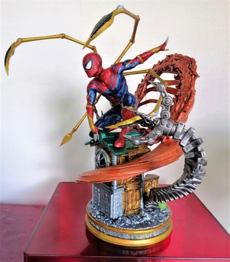 May include: A red and blue Spider-Man action figure with gold web shooters, standing on a detailed base with a metallic red and gray mechanical arm. The figure is posed in a dynamic action pose.