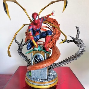 May include: A red and blue Spider-Man action figure with gold web shooters, standing on a base with a brown web and a silver mechanical arm. The figure is in a dynamic pose, with one leg raised and the other bent. The figure is made of plastic and is highly detailed.