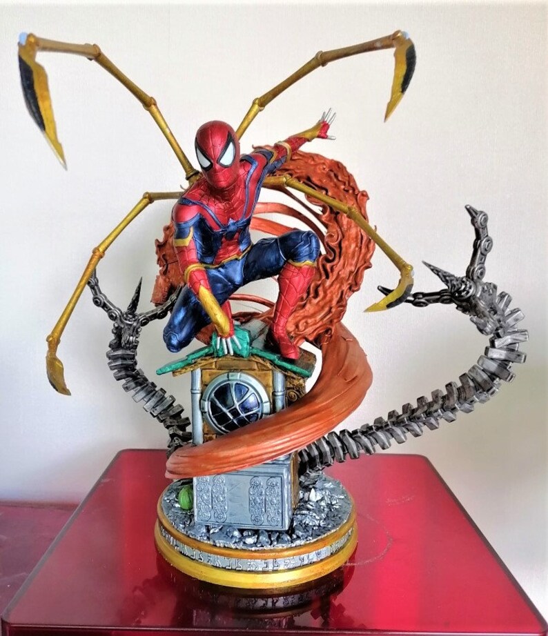 May include: A red and blue Spider-Man action figure with gold web shooters, standing on a base with a brown web and a silver mechanical arm. The figure is in a dynamic pose, with one leg raised and the other bent. The figure is made of plastic and is highly detailed.